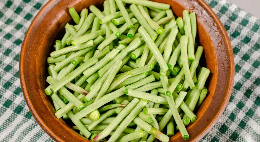 Fagiolini freschi, tagliati e pronti per essere cucinati in umido, in una ciotola di terracotta su una tovaglia a quadri verdi.