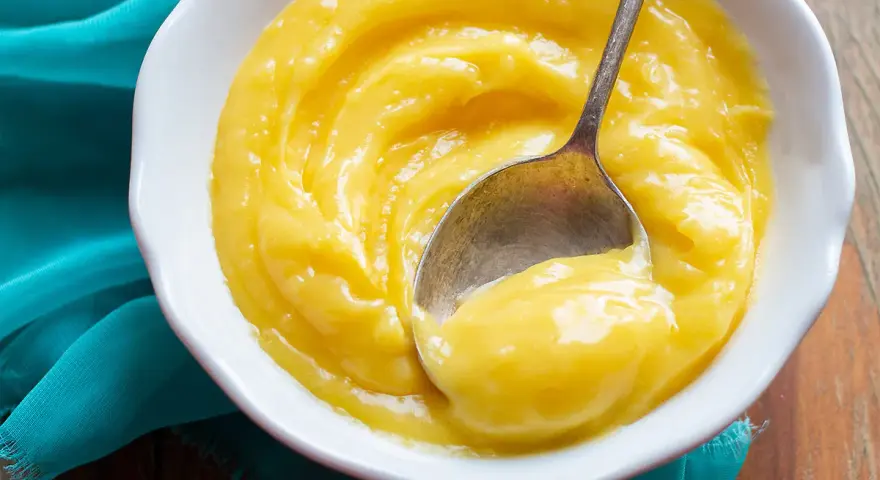 Una ciotola bianca piena di vellutata crema al limone, con un cucchiaino che ne raccoglie una porzione.