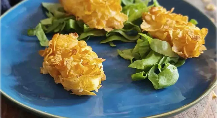 Tre croccanti polpette di patate impanate con fiocchi di mais, servite su un letto di insalata fresca su un piatto blu.