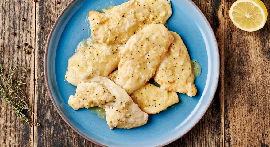 Delizioso petto di pollo in salsa cremosa, guarnito con erbe aromatiche e servito su un piatto blu. Accanto, limone e pepe nero.