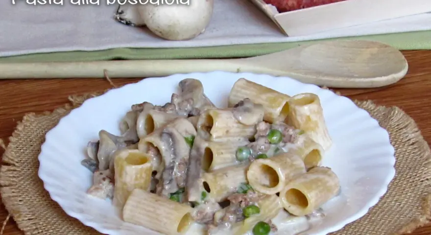Piatto di pasta alla boscaiola con funghi, piselli e salsiccia, servita su un piatto bianco bordato.