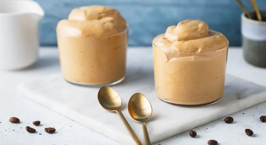Due bicchieri di cremosa schiuma di caffè, perfetti per una ricetta crema di caffè senza panna. Due cucchiaini dorati e chicchi di caffè completano la scena.