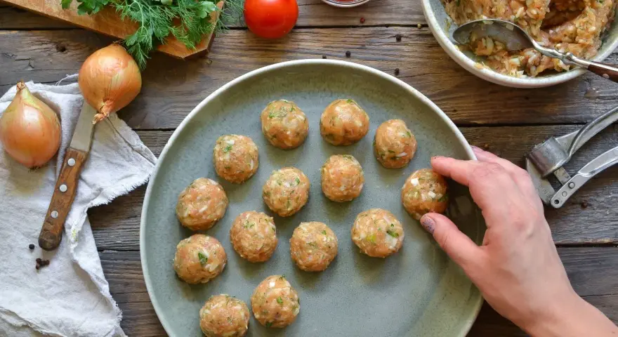 Polpette di pollo pronte per essere cucinate, disposte su un piatto grigio, con ingredienti freschi come cipolle e pomodoro sullo sfondo.