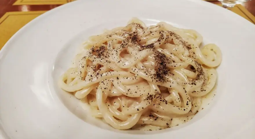 Pici cacio e pepe, un classico piatto romano, servito in un piatto bianco, con abbondante pepe nero macinato.