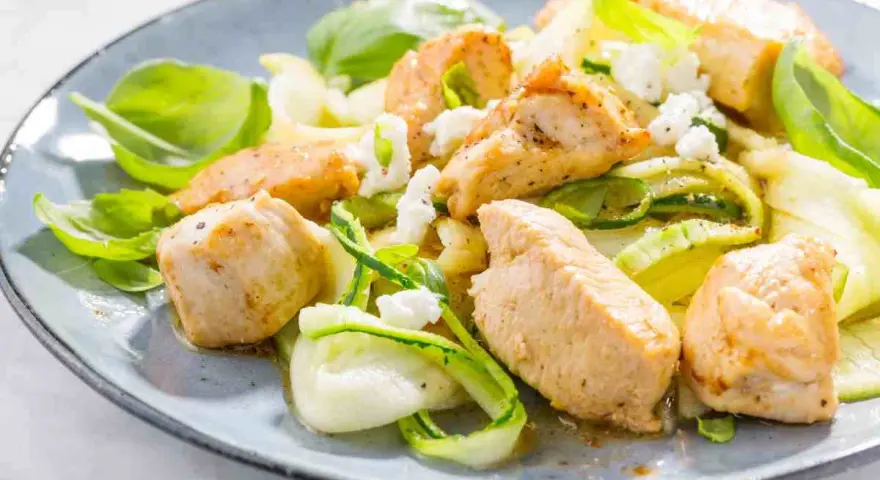 Succulento pollo e zucchine a spirale, guarnito con formaggio sbriciolato e basilico fresco, servito su un piatto blu.