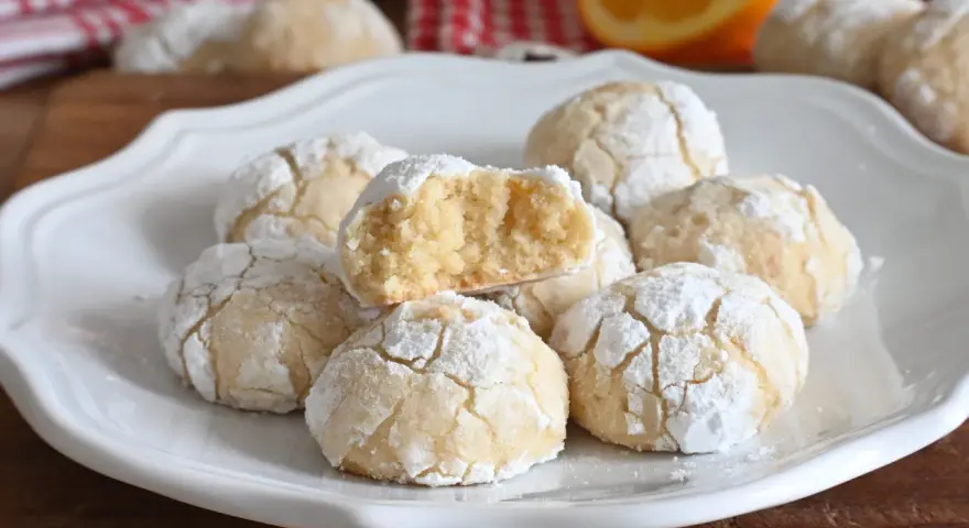 Biscotti alla ricotta soffici e profumati, cosparsi di zucchero a velo, con una fetta d'arancia sullo sfondo.