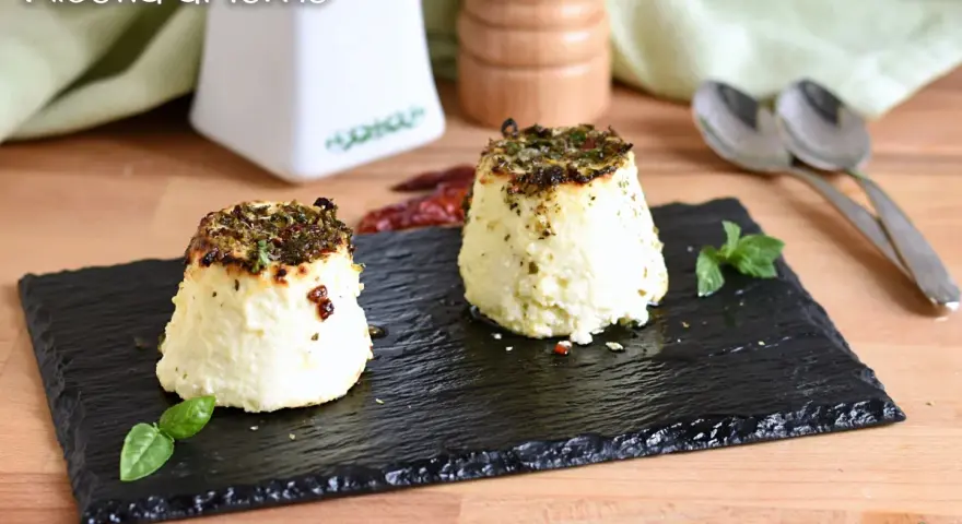 Due tortini di ricotta al forno, dorati e profumati, serviti su ardesia con basilico e peperoncino.