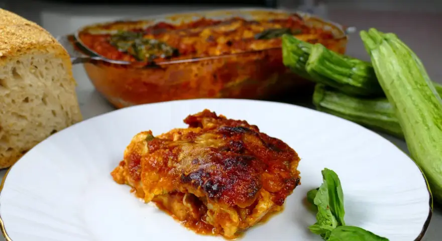 Una fetta di parmigiana di zucchine alla siciliana, dorata e succulenta, accanto a zucchine fresche e pane casereccio.