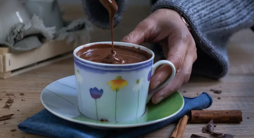 Una mano versa cioccolata fatta in casa in una tazza fiorata. Profumo di cannella e scaglie di cioccolato sul tavolo.