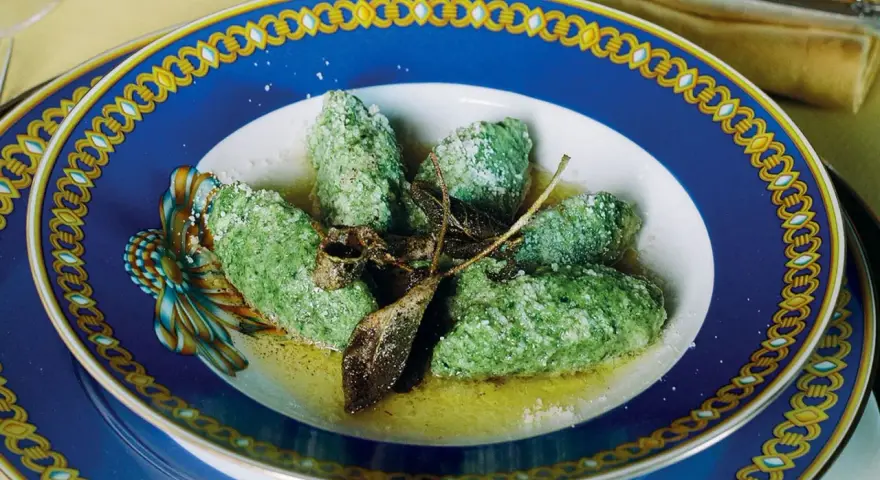 Strangolapreti alla trentina, verdi e saporiti, serviti in un piatto blu decorato con burro fuso e parmigiano.
