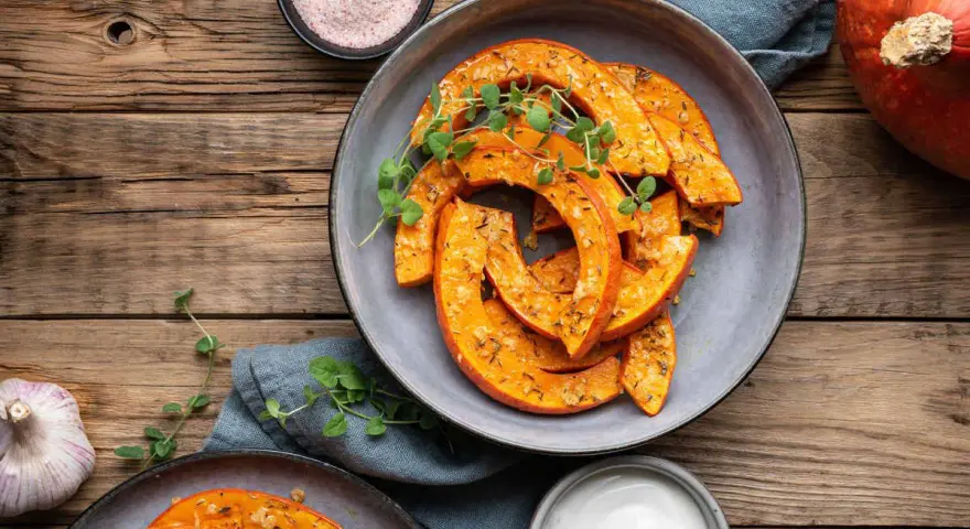 Fette di zucca in agrodolce in padella, condite con erbe aromatiche e servite su un piatto grigio, con contorno di aglio e sale rosa.
