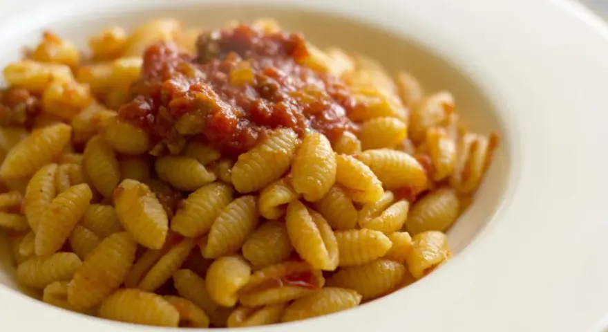 Malloreddus alla campidanese, pasta sarda con sugo di pomodoro e salsiccia, serviti in un piatto bianco.