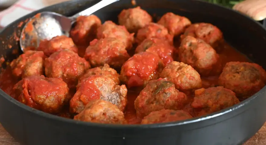 Un tegame pieno di polpette al sugo, pronte per essere gustate. Un cucchiaio d'argento è appoggiato sul bordo, invitando a servirle.