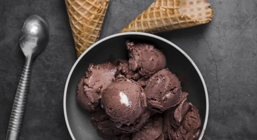 Coppa di **gelato al cioccolato** cremosa, con coni vuoti e un cucchiaio d'argento su sfondo scuro.