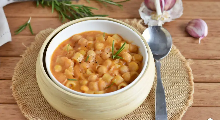 Un piatto fumante di pasta e ceci, guarnito con rosmarino fresco, su un tavolo di legno. Accanto, spicchi d'aglio e un cucchiaio.