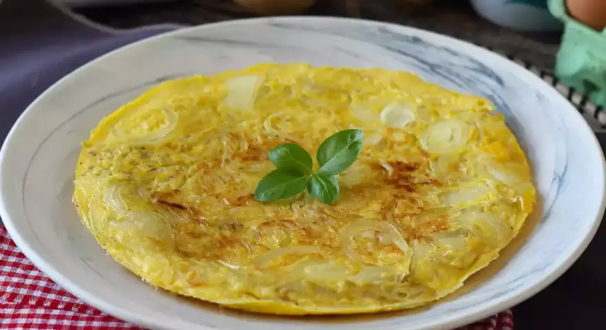 Una dorata frittata di cipolle, guarnita con basilico fresco, servita su un piatto bianco.