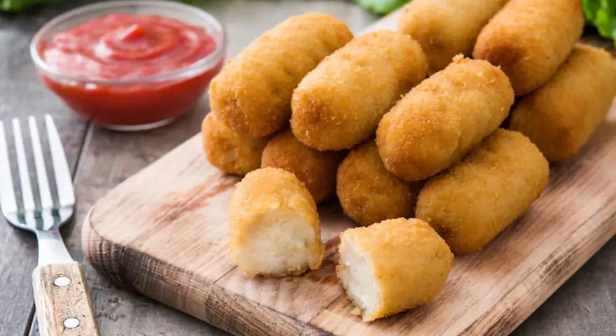 Crocchette di patate dorate e croccanti, servite su un tagliere di legno con ketchup e una forchetta.