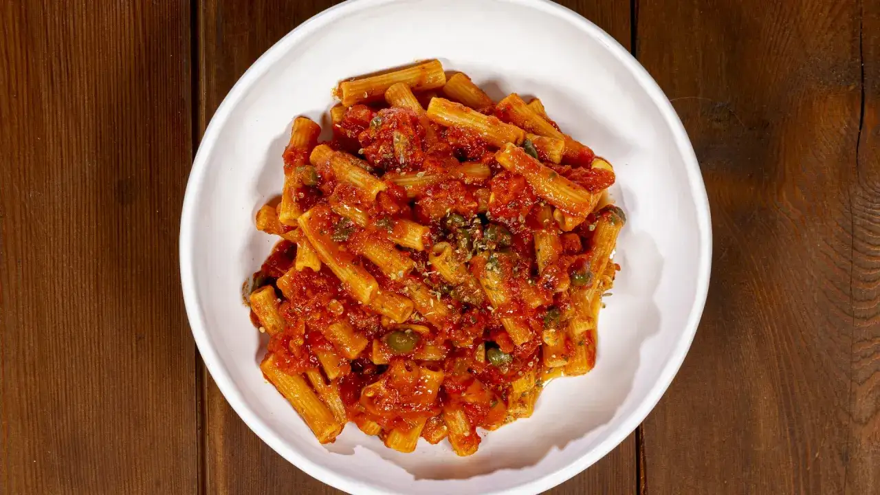 Un piatto di pasta alla pizzaiola, con rigatoni conditi con sugo di pomodoro, capperi e origano, servito in una ciotola bianca su un tavolo di legno scuro.