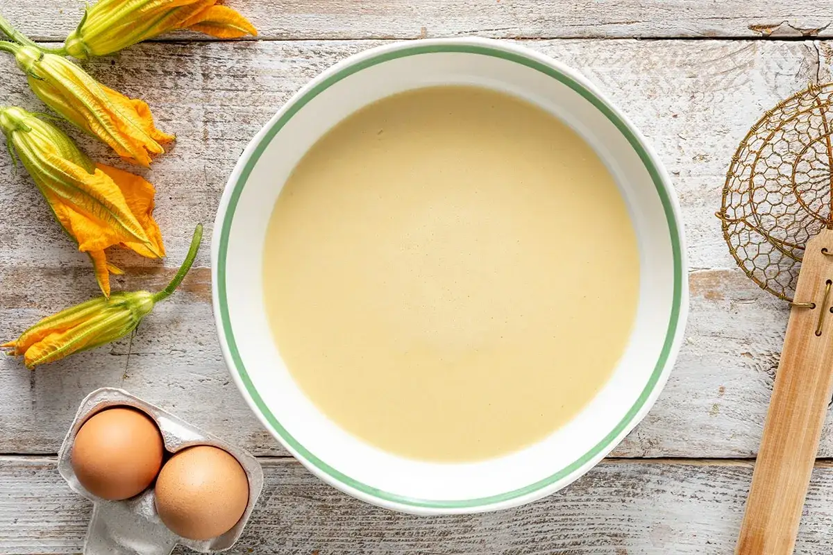 Ingredienti per i fiori di zucca in pastella: fiori freschi, uova e una ciotola di pastella dorata pronta per l'uso.
