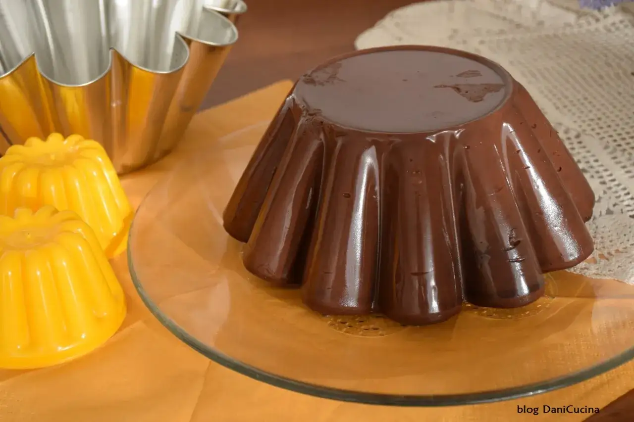 Budino fatto in casa al cioccolato, lucido e invitante, pronto per essere gustato. Accanto, due piccoli budini gialli e uno stampo metallico.