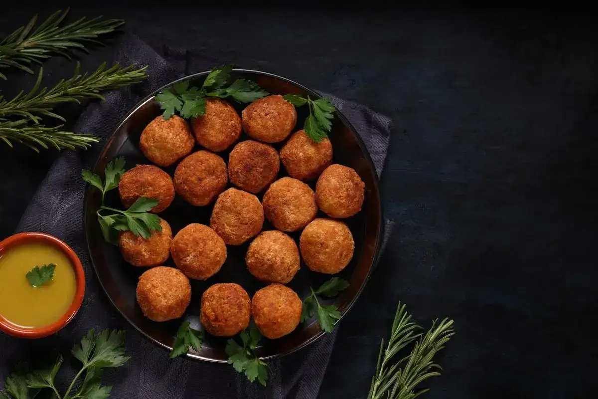 Piatto di polpette dorate con peperoni, guarnite con prezzemolo fresco e accompagnate da una ciotolina di salsa gialla.