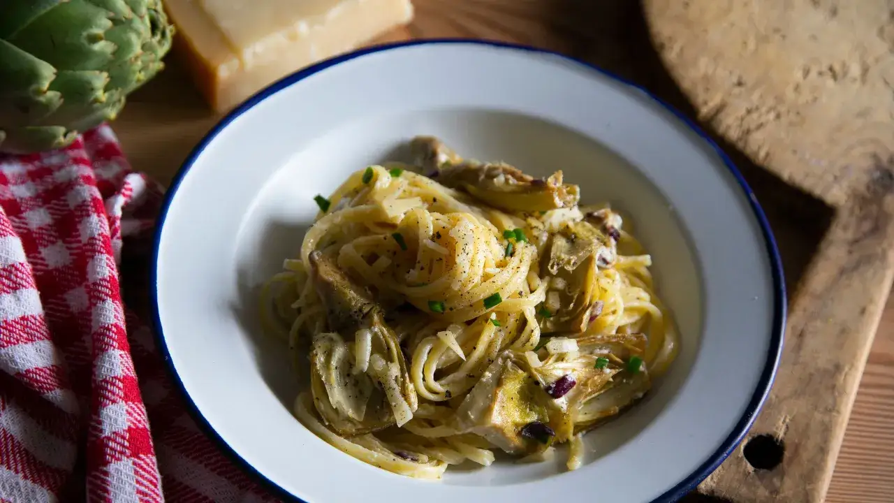 Un piatto di pasta con i carciofi cremosa, condita con parmigiano e pepe nero. Sullo sfondo, un carciofo fresco e un pezzo di formaggio.