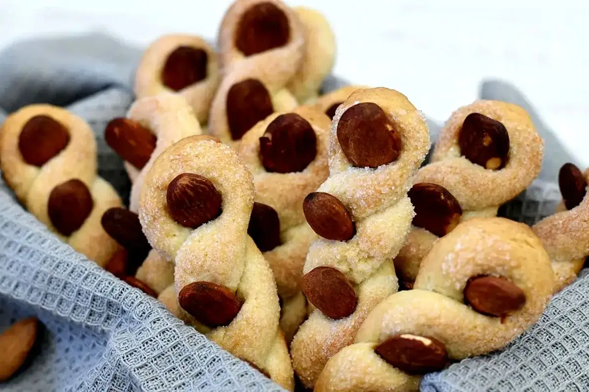 Intorchiate pugliesi ricetta originale: biscotti intrecciati con mandorle, cosparsi di zucchero, in un cestino foderato di stoffa grigia.