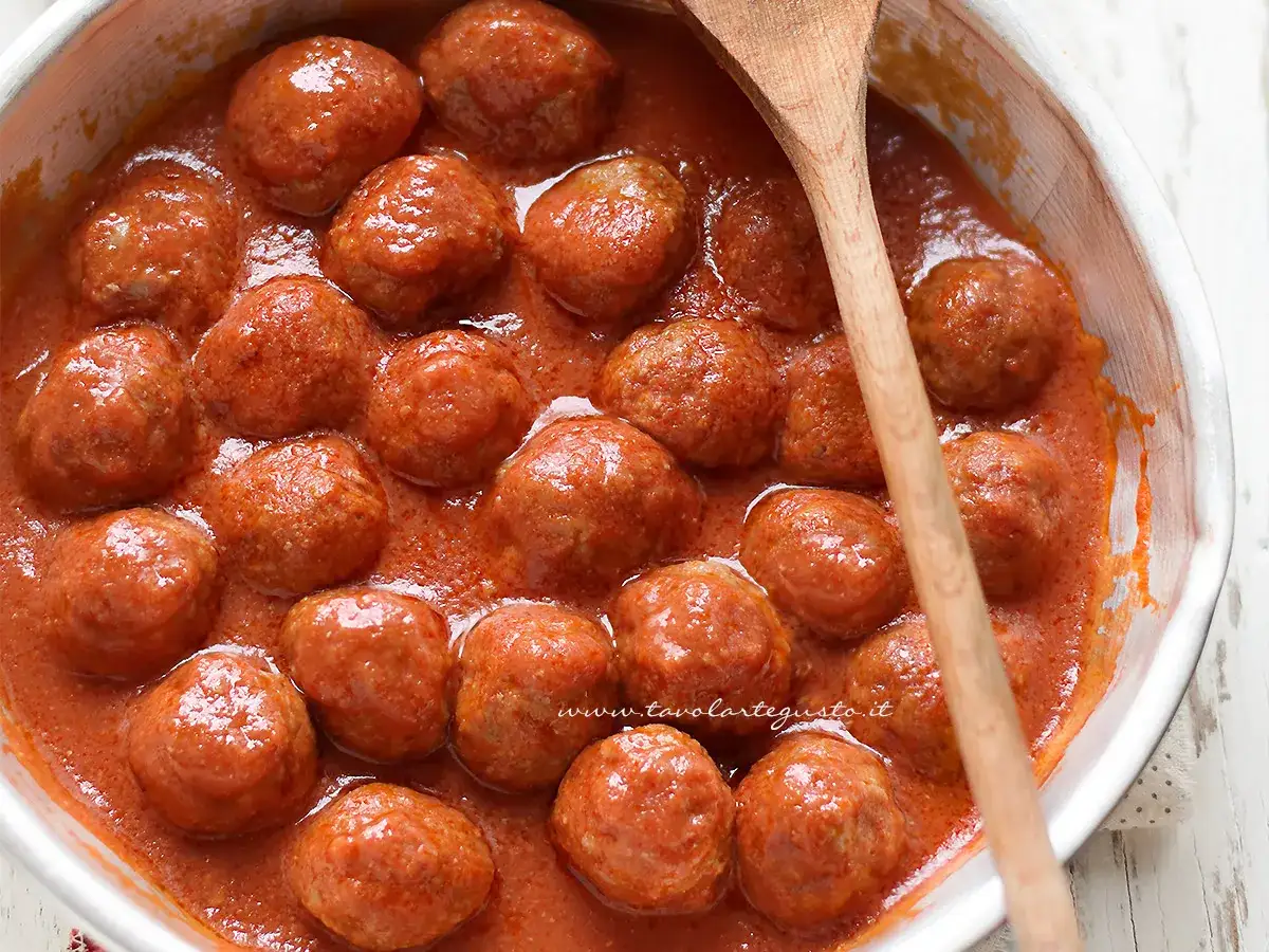 Succulente polpette di carne in un sugo rosso e denso, pronte per essere gustate. Un cucchiaio di legno attende di servirle.