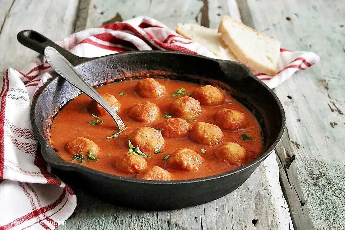 Polpette di pane al sugo in padella, un classico confortante. Accompagnate da fette di pane fresco e un tovagliolo a quadri rossi.