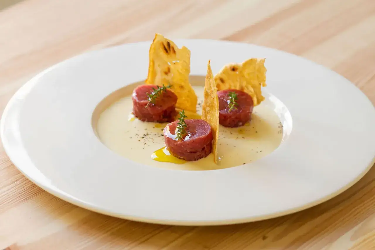 Tre porzioni di tartare di manzo, guarnite con erbe aromatiche e accompagnate da croccanti sfoglie dorate, adagiate su una crema chiara.