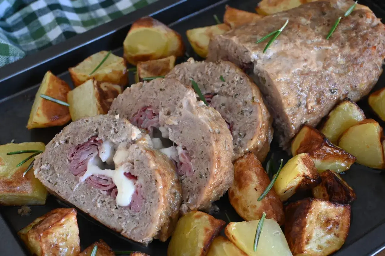 Sliced meatloaf stuffed with ham and cheese, served with roasted potatoes and rosemary on a baking sheet.