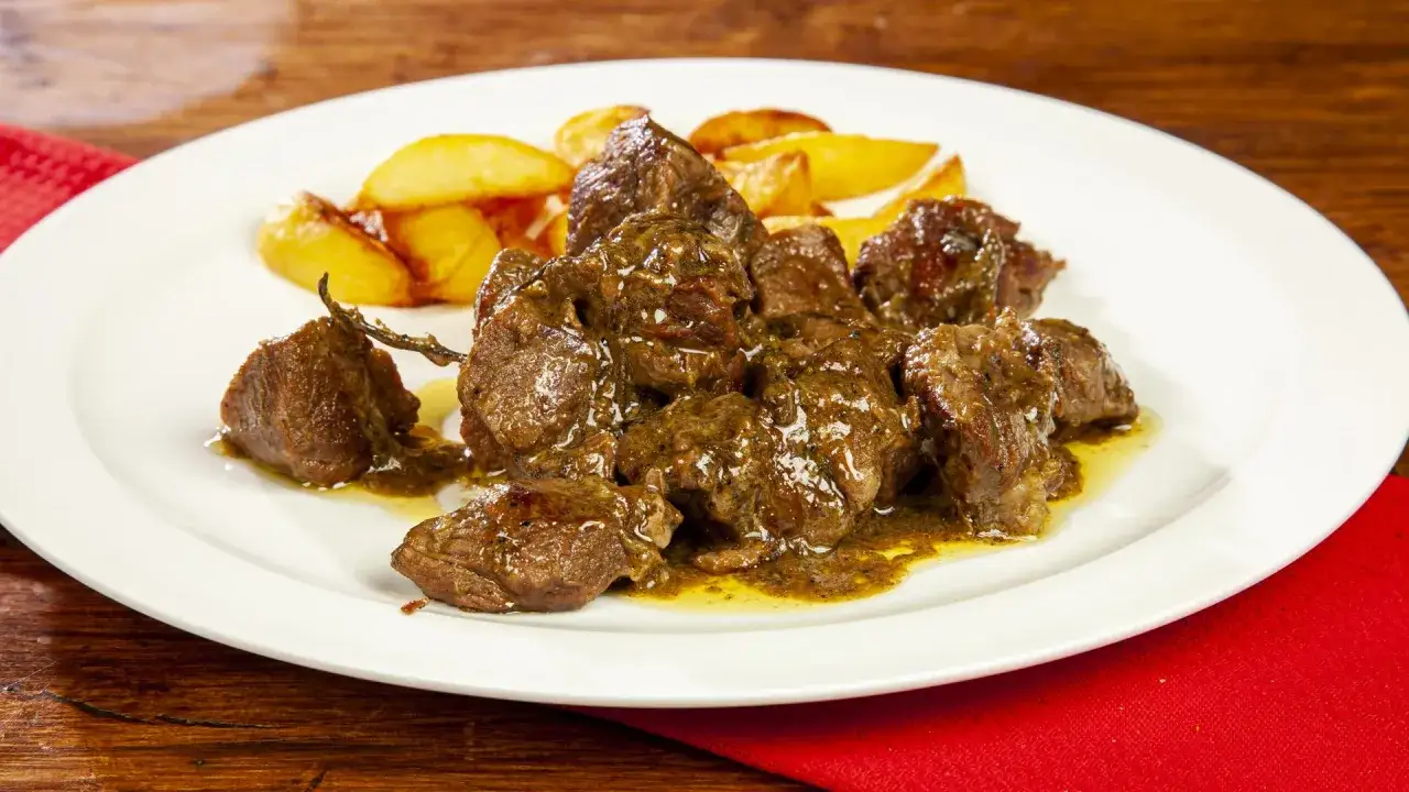 Piatto di abbacchio alla cacciatora succulento, con patate arrosto dorate, servito su un piatto bianco con tovagliolo rosso.