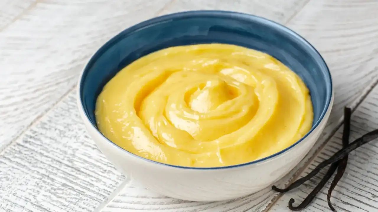 Una ciotola blu piena di vellutata crema pasticcera, con baccelli di vaniglia scuri accanto.