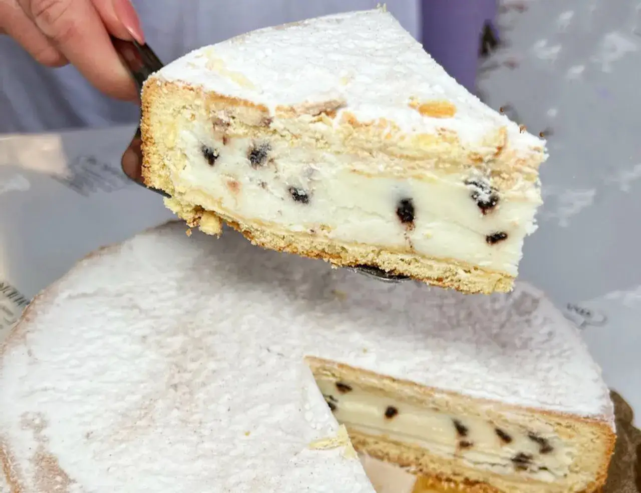 Una fetta di cassata al forno, ricca di crema e gocce di cioccolato, viene servita da una torta intera cosparsa di zucchero a velo.