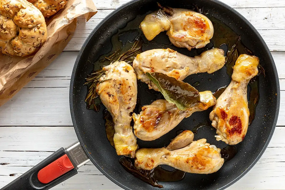 Cosce di pollo in padella, dorate e succulente, con rosmarino e alloro. Accanto, panini integrali appena sfornati.