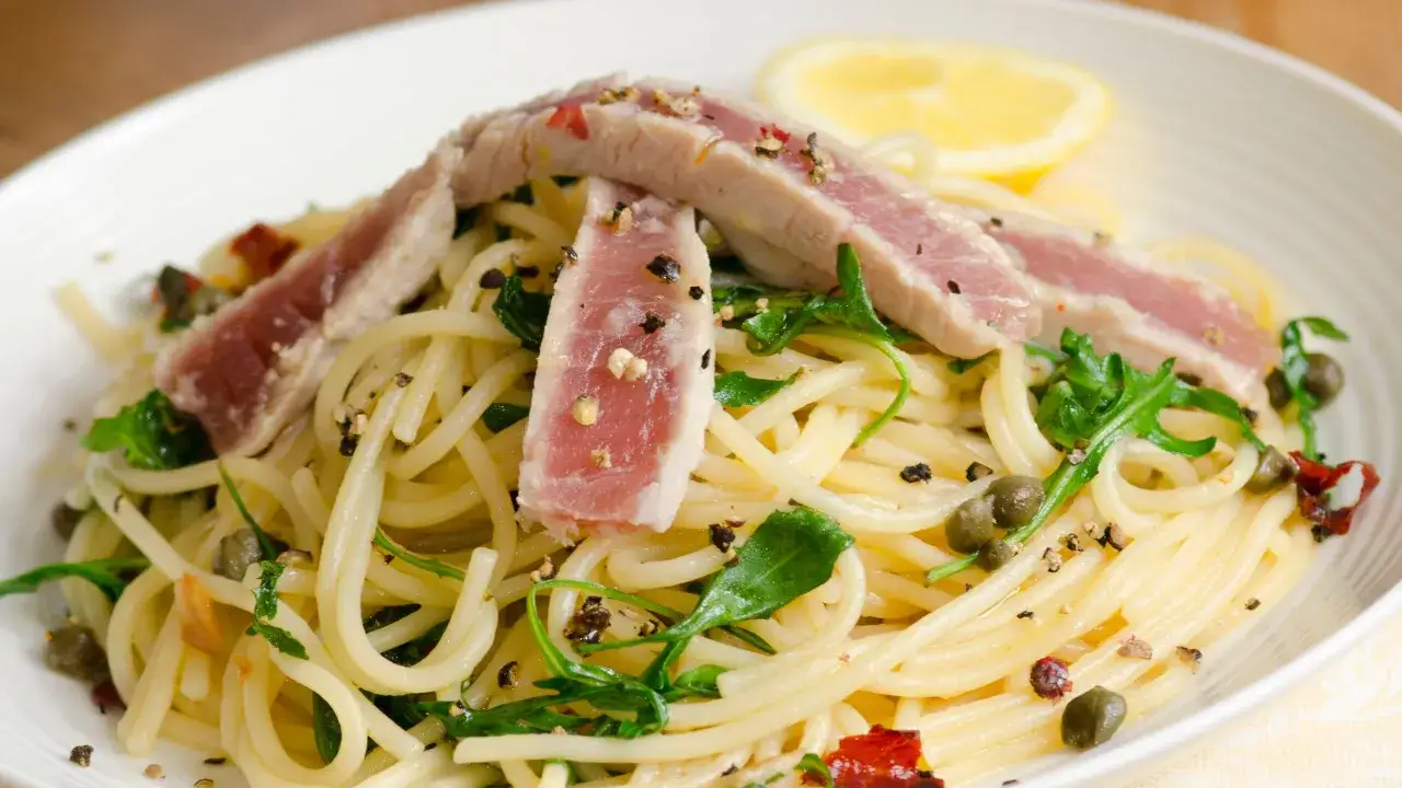 Spaghetti con tonno fresco alla siciliana, conditi con capperi, pomodori secchi, rucola e una fetta di limone.