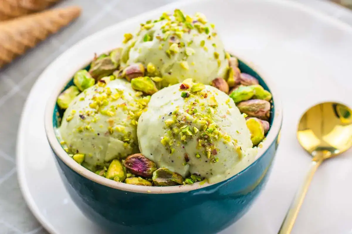 Coppa di gelato al pistacchio, guarnita con granella e pistacchi interi. Un cucchiaio dorato attende accanto.