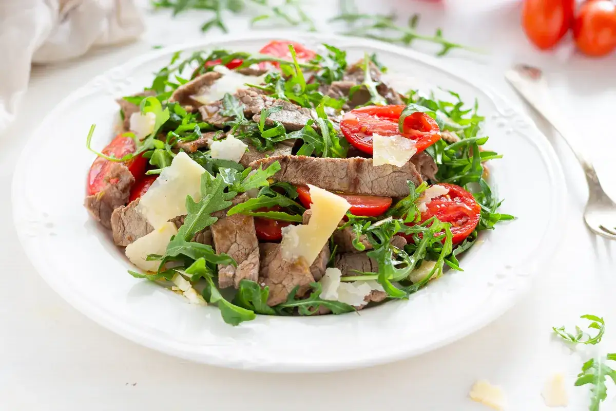 Insalata fresca con straccetti di manzo, pomodorini e scaglie di parmigiano su un piatto bianco.
