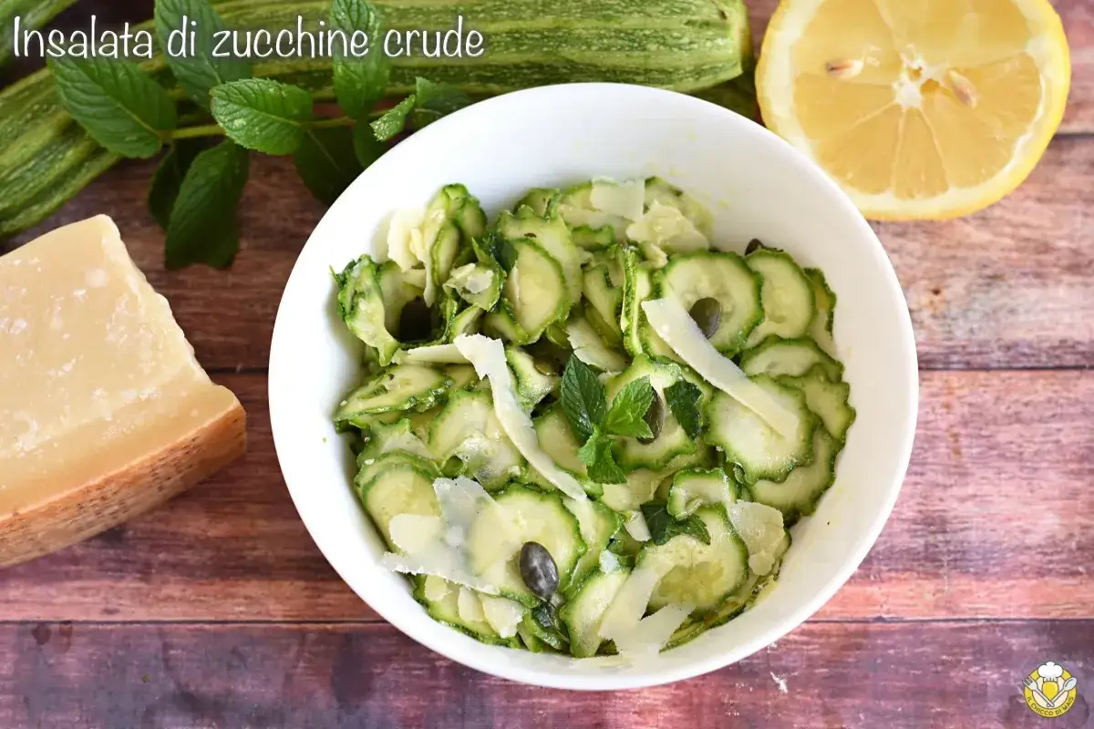 Insalata di zucchine crude, fresca e leggera, condita con scaglie di parmigiano, menta e semi di zucca. Accanto, zucchine fresche, un limone e un pezzo di parmigiano.