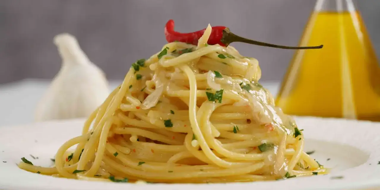 Spaghetti aglio olio e peperoncino cremosi, guarniti con un peperoncino rosso e prezzemolo. Sullo sfondo, aglio e olio d'oliva.