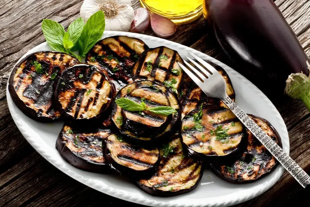 Fette di melanzane grigliate, condite con erbe aromatiche, su un piatto bianco, con una forchetta decorata.