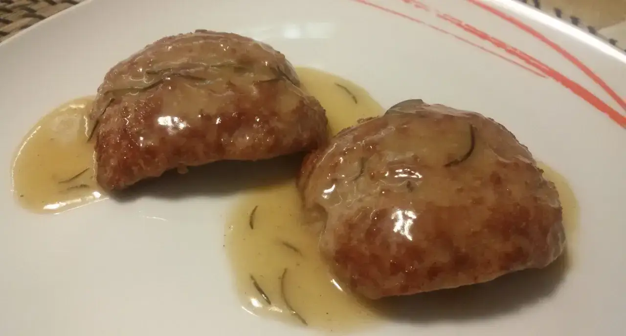 Due medaglioni di salsiccia in padella, dorati e succulenti, adagiati su un letto di sughetto aromatico con rosmarino.