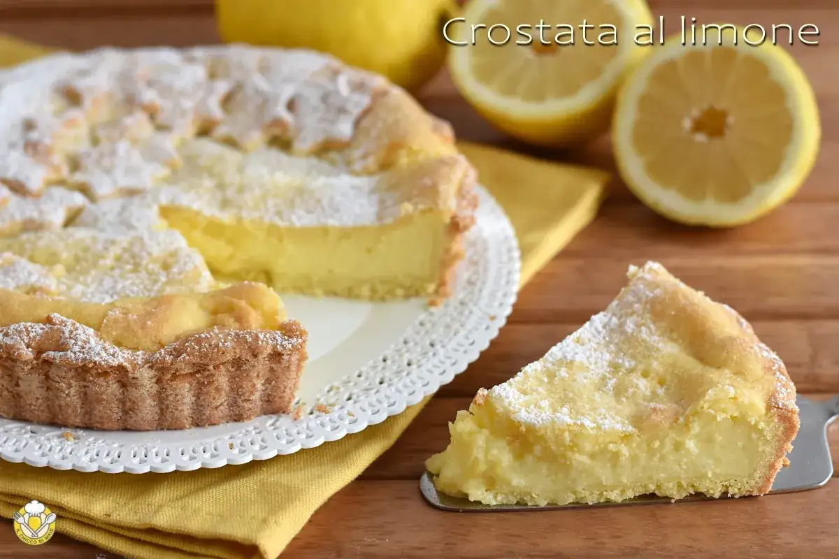 Fetta di crostata con crema al limone, cosparsa di zucchero a velo. Sullo sfondo, limoni freschi e la torta intera.