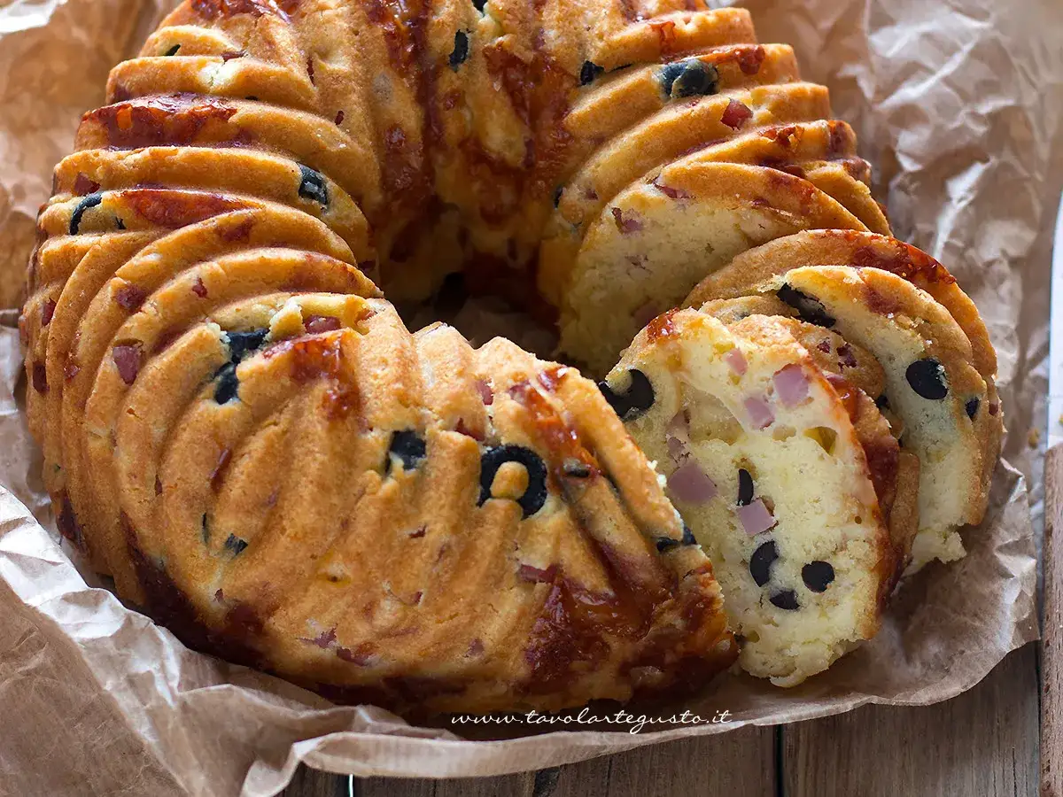 Ciambella rustica morbidissima con olive nere e cubetti di prosciutto, tagliata a fette su carta da forno.