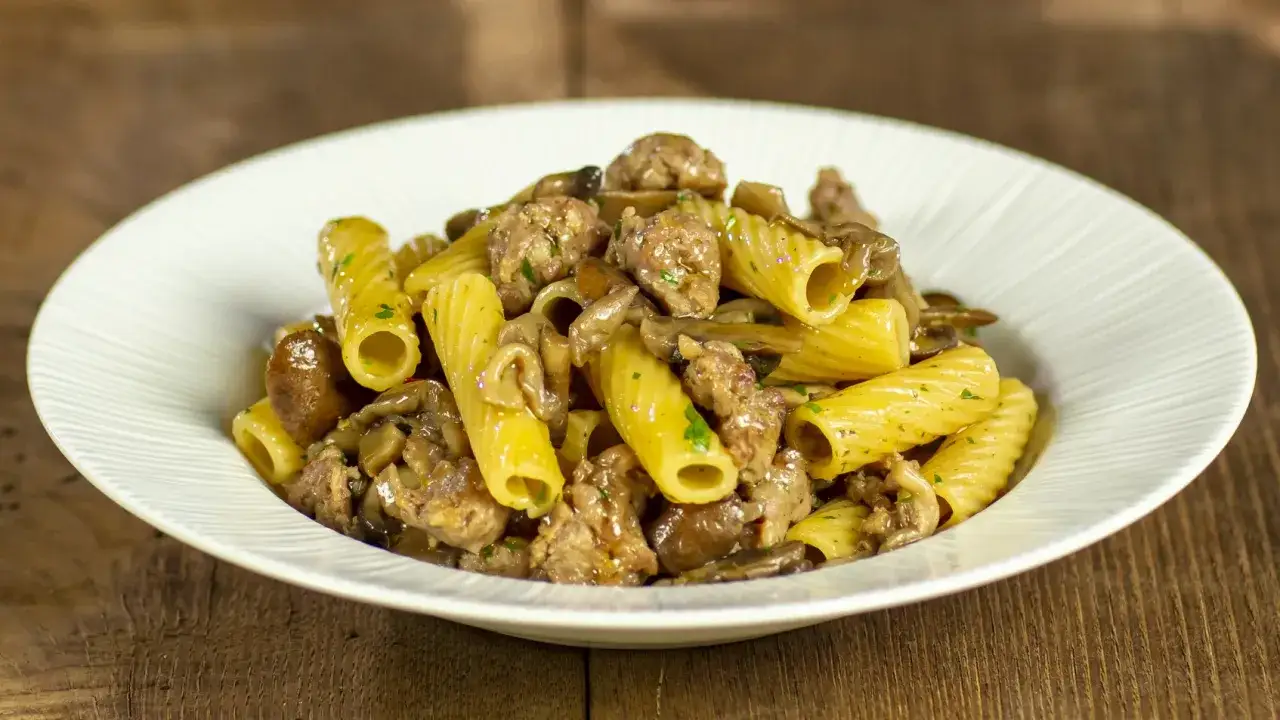 Piatto di pasta alla boscaiola con rigatoni, salsiccia e funghi, guarnito con prezzemolo tritato.