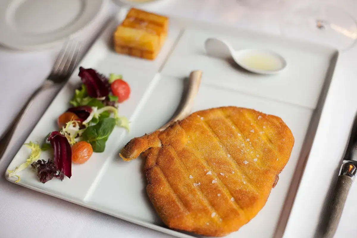Cotoletta alla milanese dorata e croccante, servita con insalata fresca, pomodorini e una piccola ciotola di limone.