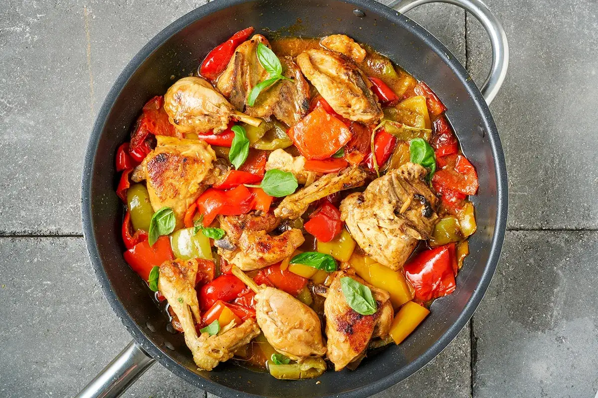 Pollo con peperoni alla siciliana in padella, con pezzi dorati di pollo e peperoni rossi e gialli, guarnito con basilico fresco.