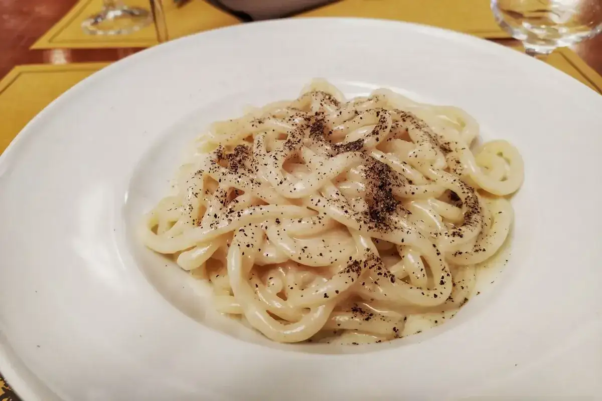 Piatto di pici cacio e pepe, pasta fatta a mano condita con pecorino romano e pepe nero macinato fresco, servita su un piatto bianco.