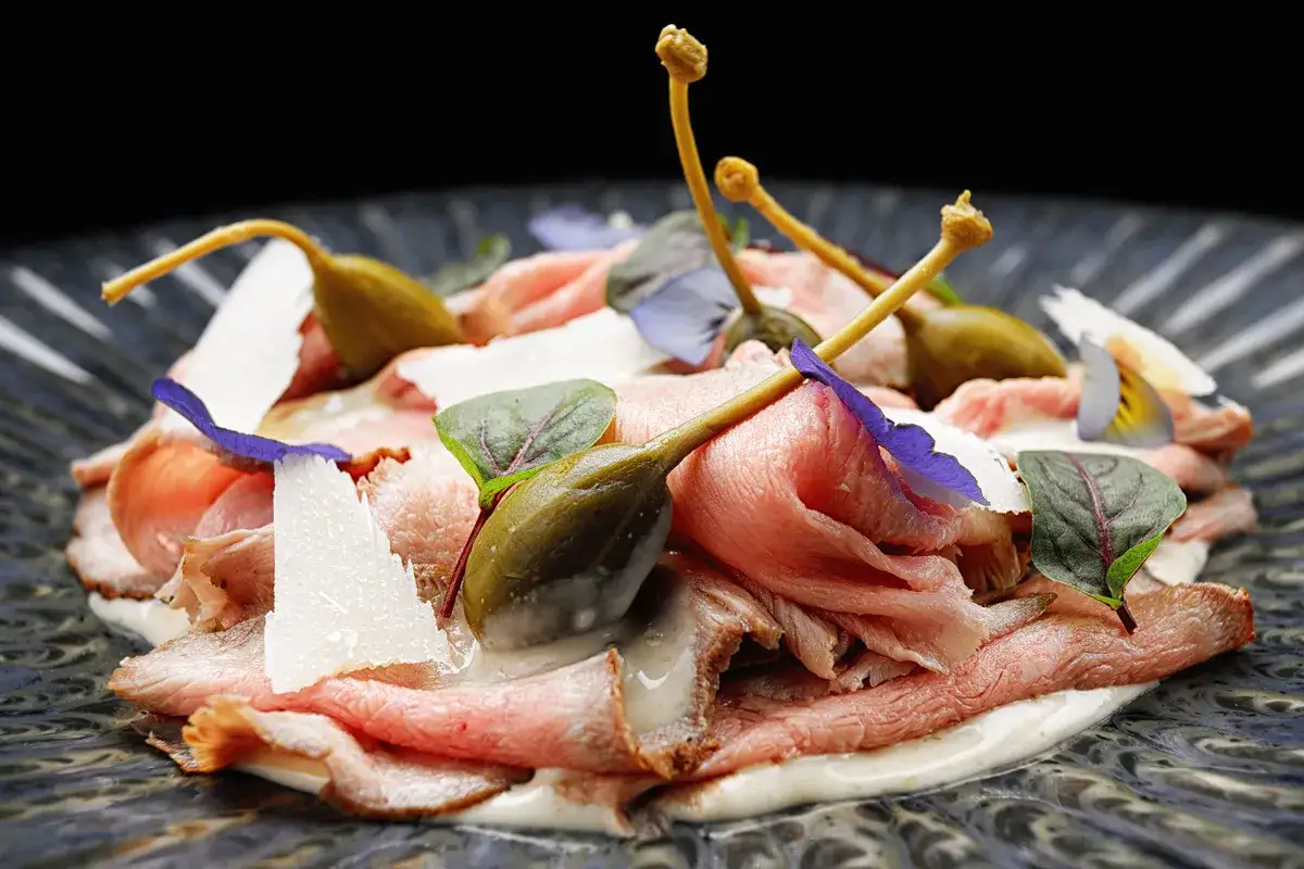 Vitello tonnato: fettine sottili di arrosto di vitello, nappate con salsa tonnata, guarnite con capperi, scaglie di parmigiano e fiori eduli.