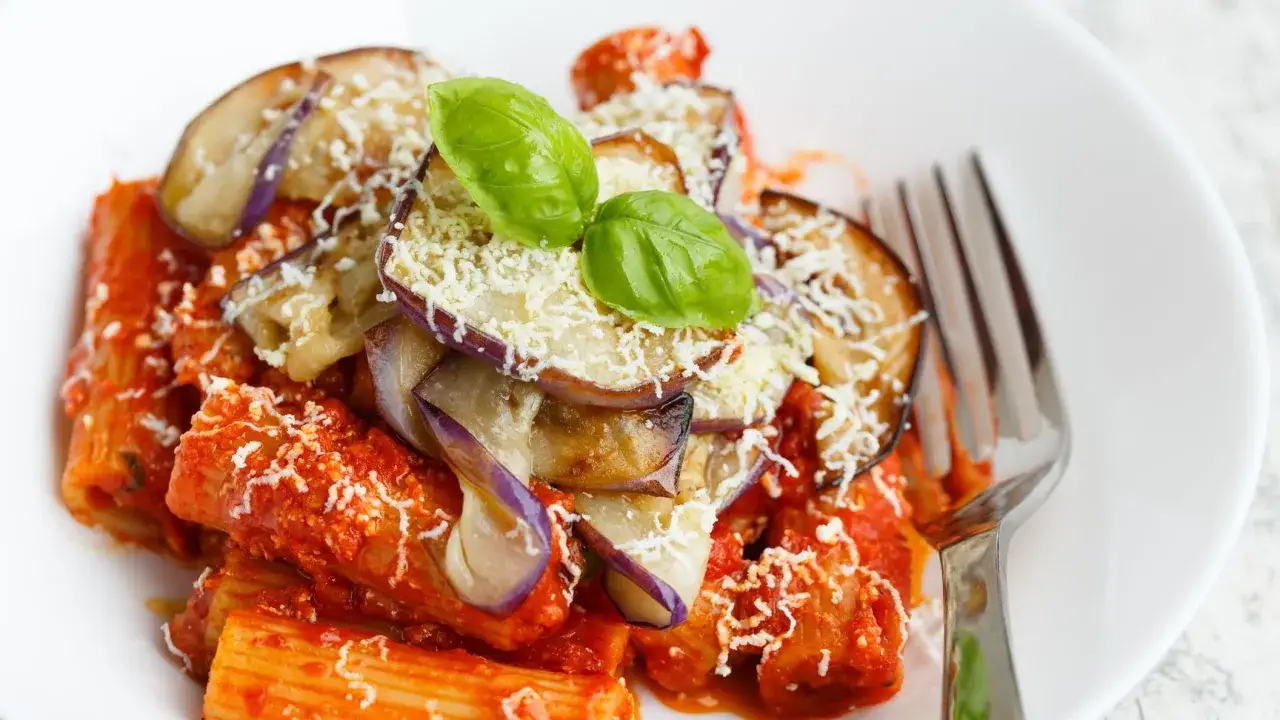 Un piatto di pasta alla norma con melanzane fritte, salsa di pomodoro e ricotta salata grattugiata, guarnito con basilico fresco.
